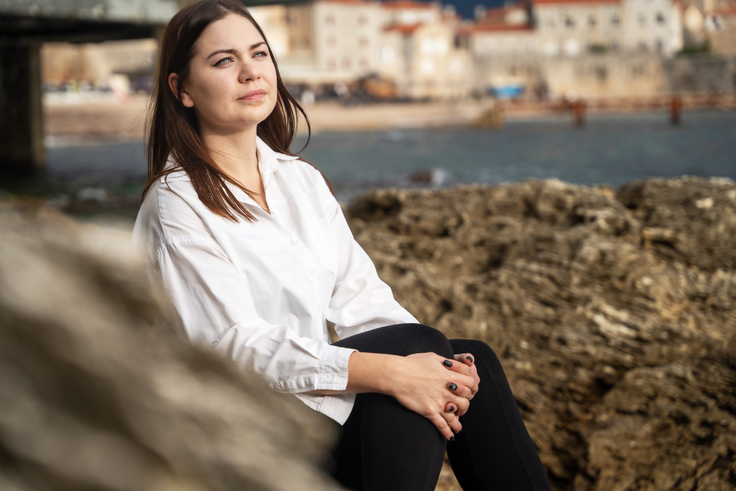 Beautiful Young Brunette Woman Sits On Stone Pier And Gazes Thou