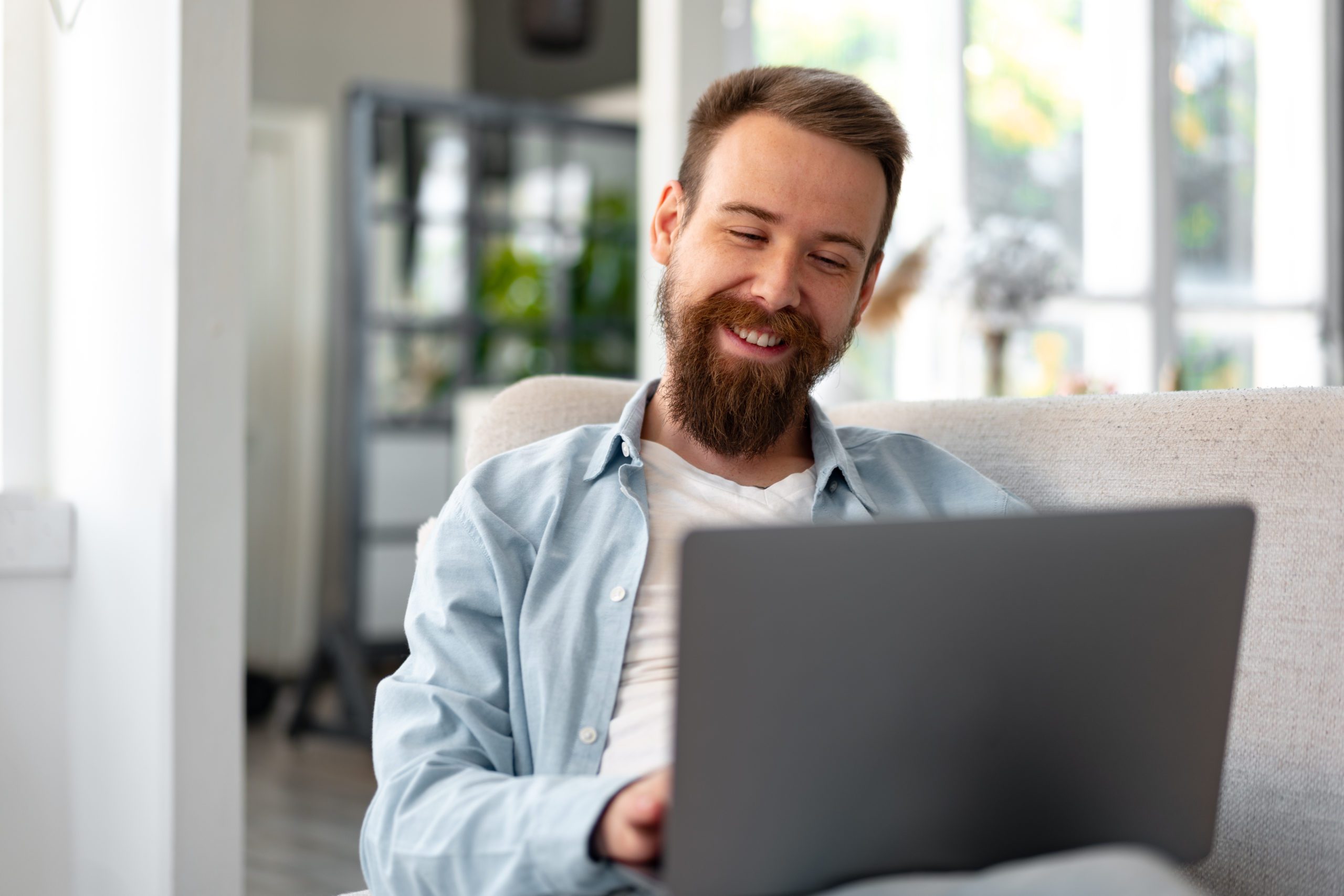 Young Bearded Man Sitting On Sofa At Home Using Laptop Computer
